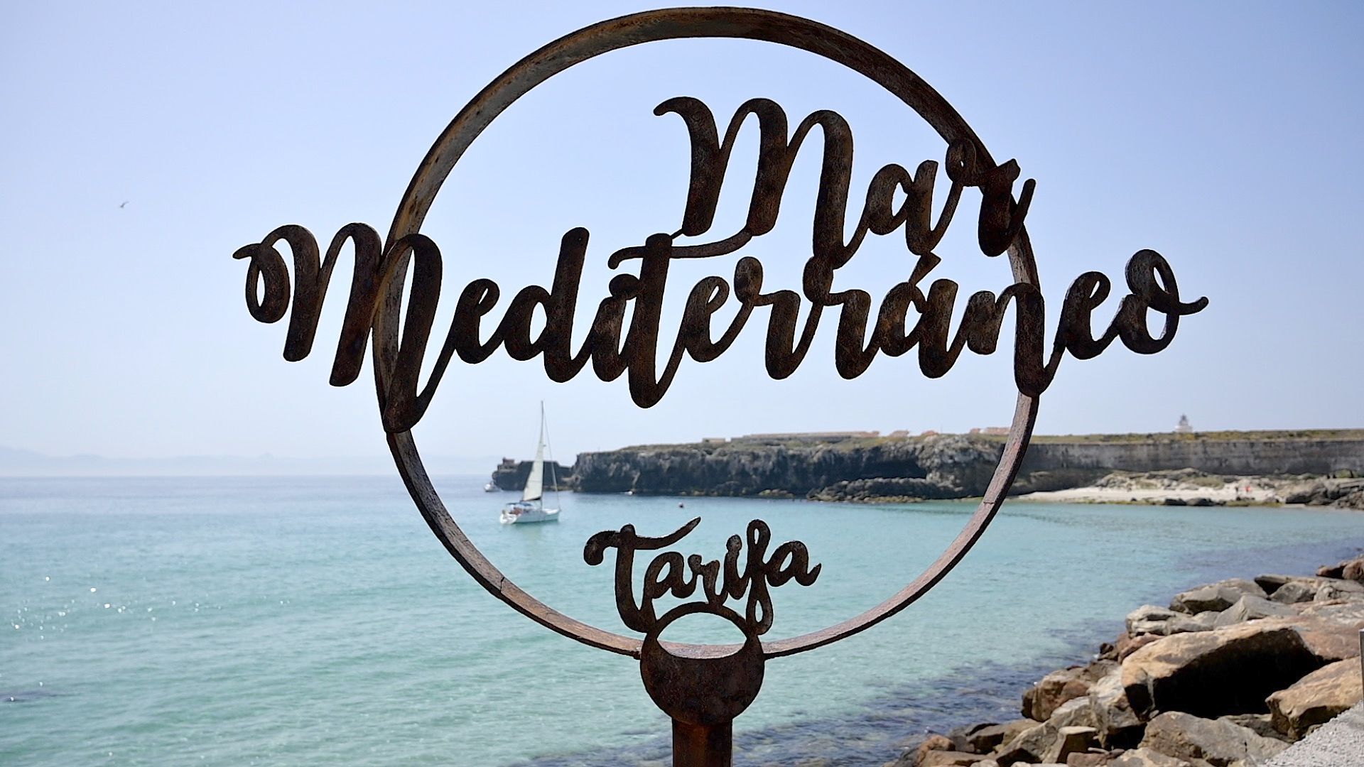 Escultura 'Mar Mediterráneo · Tarifa' frente a la cala de aguas turquesas, con un velero al fondo