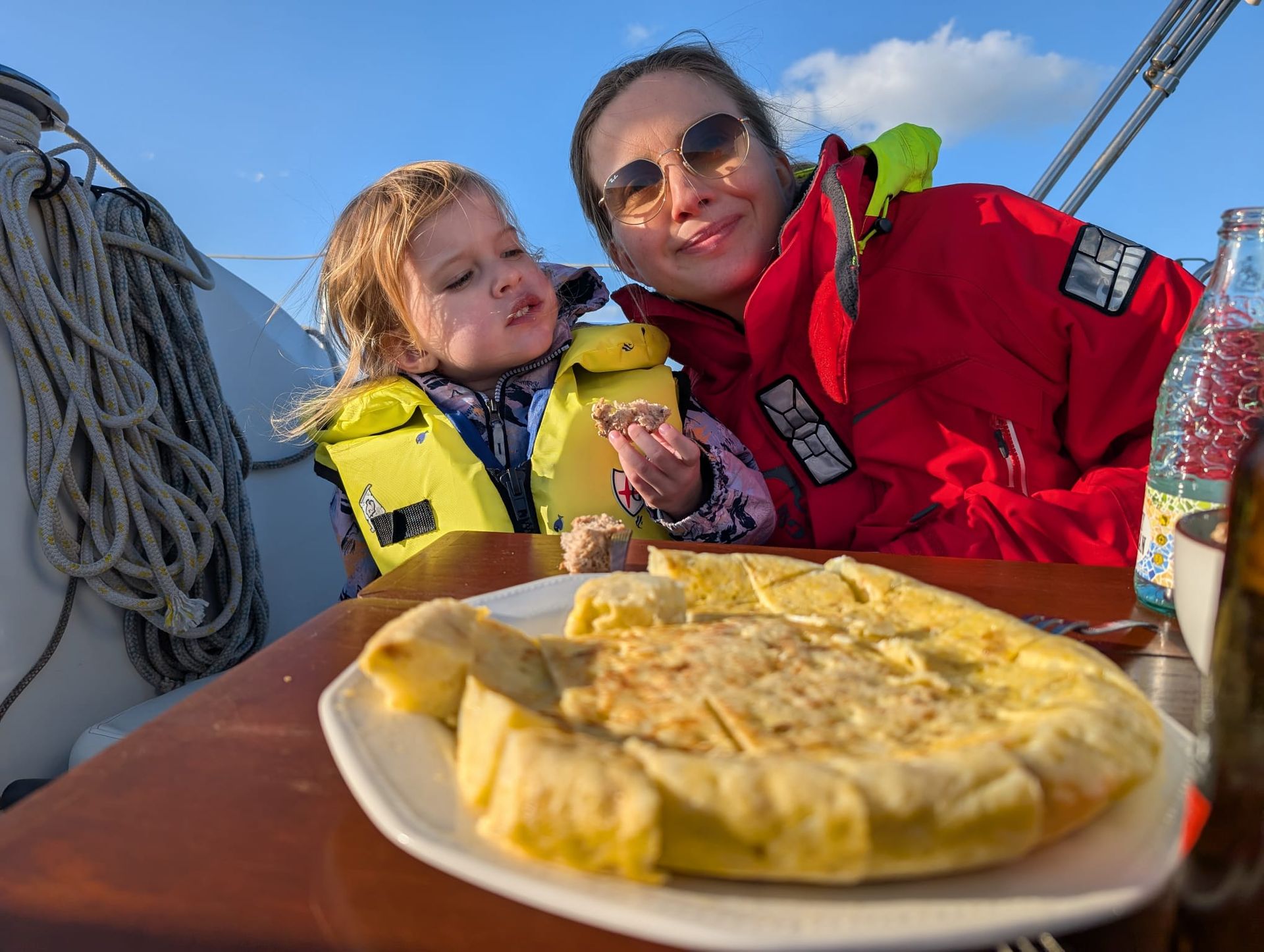 Una madre y su hija pequeña, con chalecos salvavidas, comparten tortilla casera a bordo bajo un cielo azul