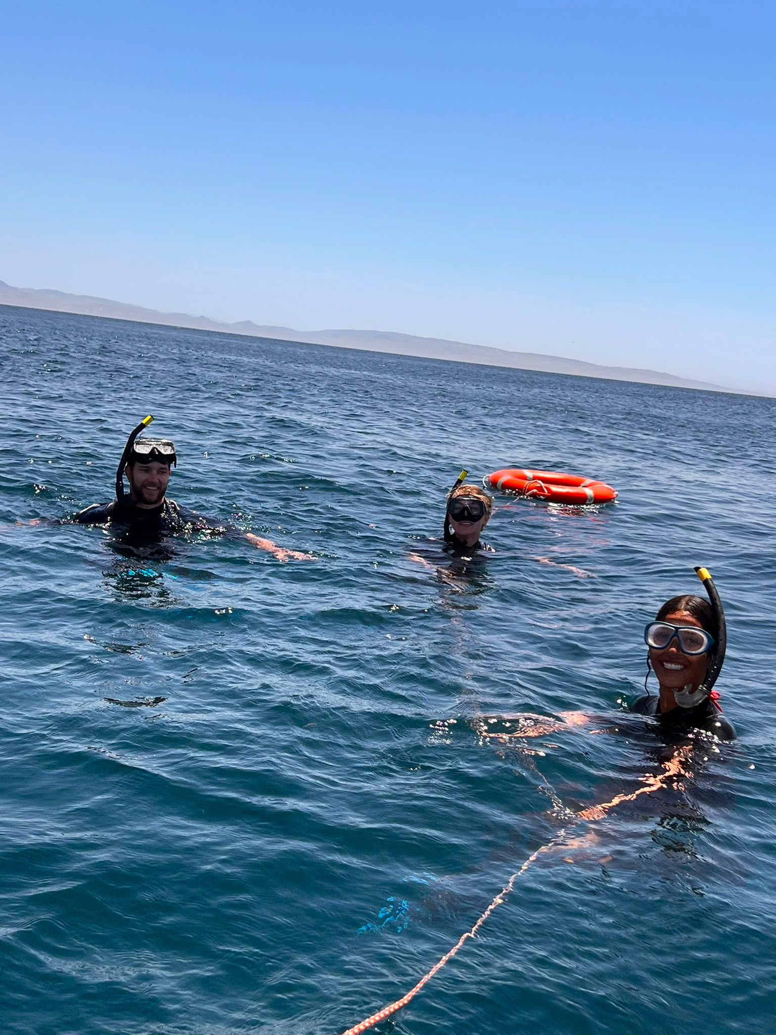 Tres pasajeros haciendo esnórquel en aguas turquesas junto a una boya, con la costa al fondo